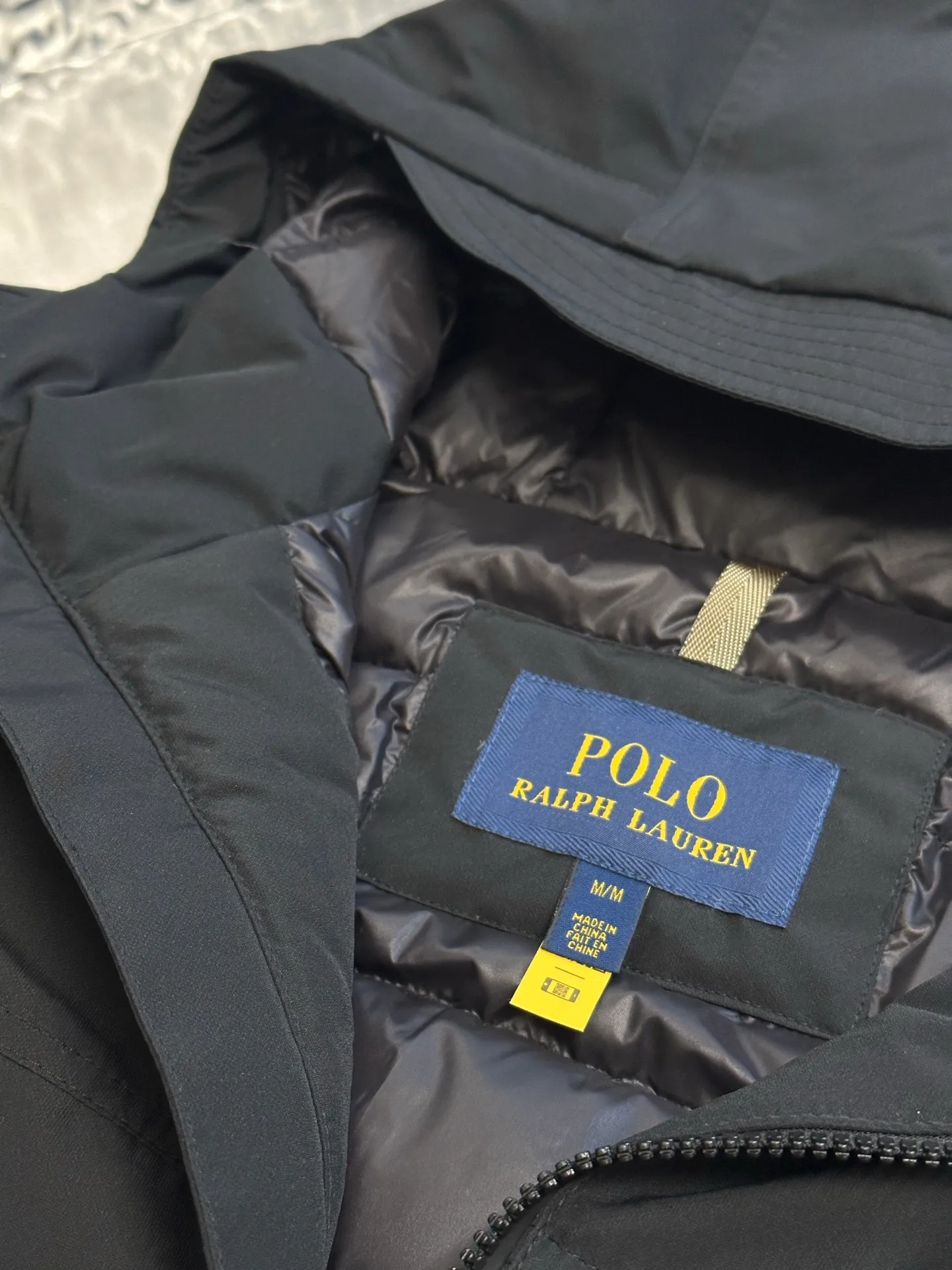 P970 Ralph Lauren logo hooded down jacket. The density of high-density nylon taffeta is about 210T-300T. The surface is smooth and slightly matte. It has excellent windproof and waterproof performance (with DWR water-repellent treatment). It has strong wrinkle resistance and is easy to care for. It is in line with the positioning of American casual "durable and practical".  The inner lining is made of 100% polyamide. The high-density and tight weaving process prevents fine lint from getting out of the gaps in the fabric. The smooth surface improves the putting on and taking off experience, and the excellent wear and tear resistance ensures damage caused by frequent use, while the light and soft texture does not affect the overall weight.  It uses 90 white duck down with a weight of 220g to ensure excellent thermal insulation performance. After strict screening and processing, the velvet is not easily drilled, ensuring durability. The classic jacket has a slightly loose fit, with slightly dropped shoulder lines (the shoulder width is 1-2cm wider than the standard fit). The length reaches the hips, and the sleeves are slightly tapered straight sleeves, taking into account freedom of movement and casual style.  The adjustable Velcro on the cuffs allows you to freely adjust the tightness according to the thickness of your wrist and personal preferences, effectively preventing cold wind from intruding, and improving the warmth performance. Two zippered pockets on the front are convenient for placing frequently used items, and the zipper design ensures that items are not easily lost. A built-in pocket on the left side of the chest provides a safe storage space for some small private items.  The drawstring design at the hem allows you to adjust the shape of the jacket according to your body shape and dressing needs. Whether it is loose and casual or slim and neat, you can easily control it.  Polo horse logo on the left chest: flat satin stitch embroidery (satin embroidery), using 100% polyester high color fastness embroidery thread (the color number is consistent with the brand's standard red color card), the stitch density is 12-15 stitches/cm, the outline of the horse logo is clear, and the details of the rider and horse are burr-free. Precise positioning through the template (12cm from the bottom of the collar and 8cm from the placket) ensures symmetry and centering.  size：s m l xl